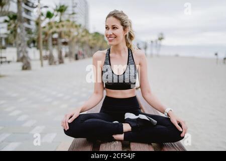 Atleta concentrato in abbigliamento e sneakers attivi alla moda seduto su panchina poggiata con traversata che guarda via con il mare su sfondo sfocato Foto Stock