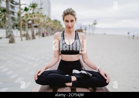 Atleta concentrato in abbigliamento e sneakers attivi alla moda seduto sul banco di appoggio con la croce gamba che guarda la fotocamera con mare su sfondo sfocato Foto Stock