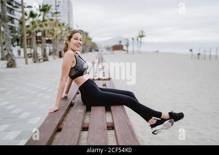 Vista laterale dell'atleta concentrato in abbigliamento attivo alla moda e le sneakers che si siedono sulla panca che riposano guardando la macchina fotografica con mare su sfondo sfocato Foto Stock