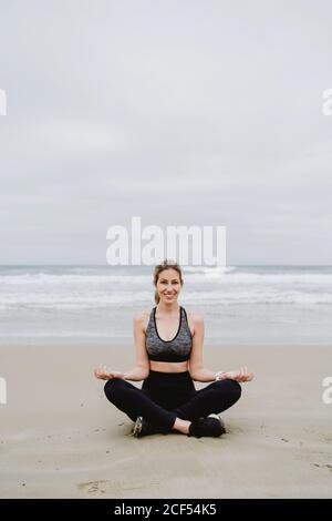 Giovane donna slim con parte superiore nera e leggings seduta su lotus posizione mentre meditating alla spiaggia Foto Stock