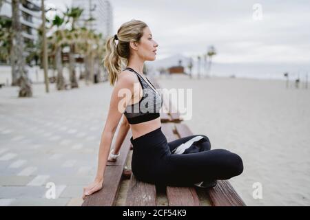 Vista laterale dell'atleta concentrato in abbigliamento attivo alla moda e le sneakers che si siedono sulla panca poggiano con l'aspetto trasversale a zampe via con il mare su sfondo sfocato Foto Stock