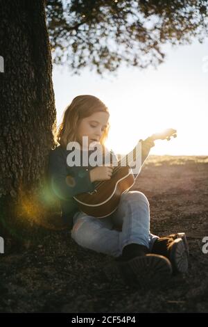 Adorabile ragazza seria in abbigliamento casual che suona la chitarra ukulele mentre si siede vicino albero in estate soleggiato giorno in campagna Foto Stock