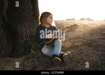 Adorabile ragazza seria in abbigliamento casual che suona la chitarra ukulele mentre si siede vicino albero in estate soleggiato giorno in campagna Foto Stock