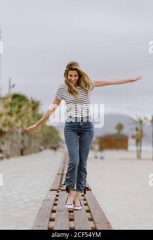 Felice donna allegra in abiti casual a piedi con le braccia sparse su una panchina di legno e godendo di una giornata estiva sul lungomare della città Foto Stock