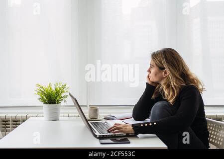 Vista laterale ad alto angolo di una donna specializzata in un ambiente casual indossare i documenti elettronici sul computer portatile mentre si è seduti a. tavolo vicino alla finestra in camera moderna a casa Foto Stock