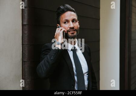 Adulto bello elegante uomo d'affari in abito formale guardando via e. parlare sul telefono cellulare vicino alla parete Foto Stock