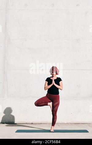 Atleta femminile sicura e barefoted in abbigliamento sportivo meditando mentre si sta in piedi vrikshasana posizione con le mani chiuse al petto in namaste su addestramento di tappetino sportivo da solo su strada contro parete di cemento alla luce del sole Foto Stock