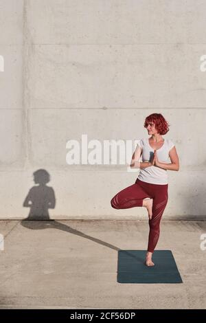 Atleta femminile sicura e barefoted in abbigliamento sportivo meditando mentre si sta in piedi vrikshasana posizione con le mani chiuse al petto in namaste su addestramento di tappetino sportivo da solo su strada contro parete di cemento alla luce del sole Foto Stock