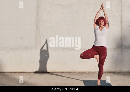 Atleta femminile sicura e barefoted in abbigliamento sportivo meditando mentre si sta in piedi vrikshasana posizione con braccia sollevate e namaste su tappetino sportivo addestramento da solo su strada contro muro di cemento in luce solare Foto Stock