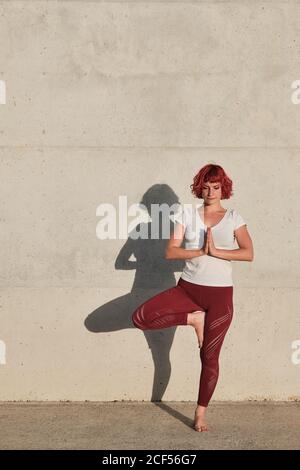 Atleta femminile sicura e barefoted in abbigliamento sportivo meditando mentre si sta in piedi vrikshasana posizione con le mani chiuse al petto in namaste su addestramento di tappetino sportivo da solo su strada contro parete di cemento alla luce del sole Foto Stock