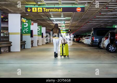 Giovane positivo appena arrivato signora in abiti casual con lungo i capelli camminano nel parcheggio e trasportano la valigia gialla nell'aeroporto edificio Foto Stock