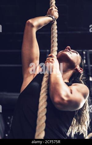 Da sotto muscoloso Donna arrampicata corda e guardando su sfondo nero Foto Stock