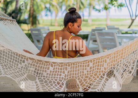 Vista posteriore di Woman felice in costume da bagno seduto in amaca e guardando giù al mare esotico in Costa Rica Foto Stock