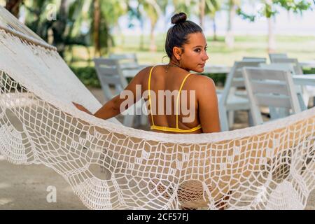 Vista posteriore di Woman entusiasta in costume da bagno seduto in amaca e guardando via il mare esotico in Costa Rica Foto Stock