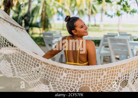 Vista posteriore di Woman entusiasta in costume da bagno seduto in amaca e guardando via il mare esotico in Costa Rica Foto Stock