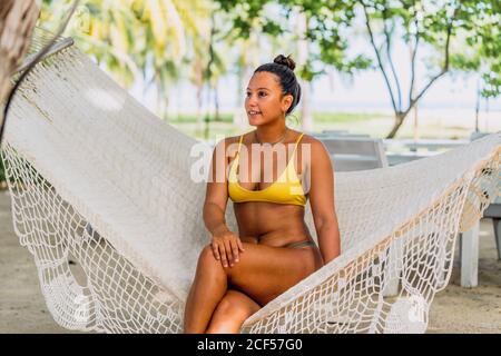 Donna felice in costume da bagno seduta in amaca e guardando lontano esotico mare in Costa Rica Foto Stock