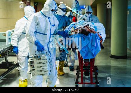 Gruppo di medici professionisti irriconoscibili che indossano maschere protettive e uniformi trasporto di pazienti con infezione virale attraverso sala in clinica a. sala operatoria Foto Stock