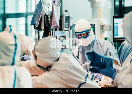 Gruppo di medici professionisti che indossano maschere protettive e tute in piedi vicino al tavolo operatorio con attrezzatura e preparazione per il funzionamento in clinica moderna Foto Stock