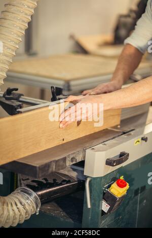 Falegname esperto che taglia un pezzo di legno nella sua officina di legno, usando una sega circolare, e indossando manichette di sicurezza e cuffie, con un altro macchinatore Foto Stock