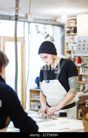 Sorridente artigiano seduto nel suo studio di lavorazione del legno per un ritratto, con mensole di diversi pezzi di legno, dove lavora come falegname Foto Stock