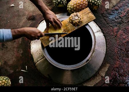 Da sopra raccolto uomo adulto tagliando ananas con coltello su parte superiore del serbatoio in metallo nella luce del giorno Foto Stock