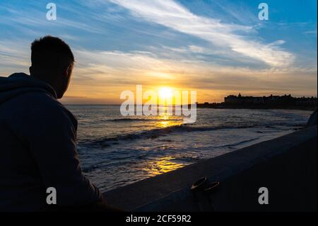 Giovane maschio sedette sulla parete guardando il tramonto a porthcawl galles uk, concentrarsi su sfondo maschile, blured Foto Stock