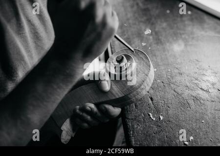 Angolo alto bianco e nero di vortice da intaglio artigiano senza volto di testa di violino mentre lavora con il dettaglio di legno usando la punta al tavolo in officina Foto Stock