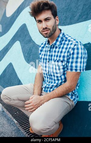 Giovane maschio allegro in camicia casual e sneakers squatting e guardando la macchina fotografica con parete dipinta sullo sfondo Foto Stock