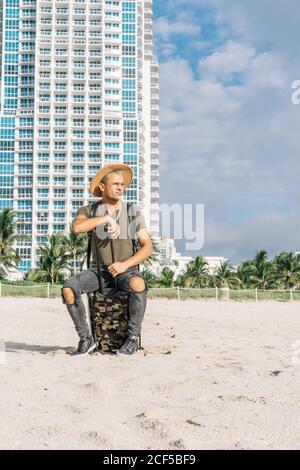 Un uomo attraente che si siede sul suo bagaglio da trasporto guardando via nella spiaggia sud di Miami. Uomo attraente seduto sul suo bagaglio da trasporto che indossa un cappello, jeans strappati e una t-shirt. Foto Stock