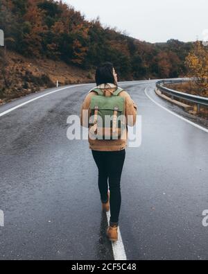 Vista posteriore di giovane escursionista attivo in abito casual con zaino che cammina su strada asfaltata umida vuota che conduce attraverso foresta colorata in autunno giorno Foto Stock
