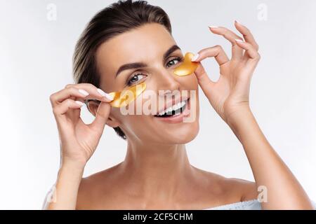 Donna felice che applica le macchie dorate di idrogel ialuronico sotto gli occhi, isolato su sfondo bianco. Cura della pelle, trattamento di bellezza per ridurre la puffiness Foto Stock