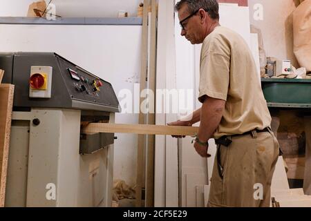 Vista laterale ad angolo basso di un artigiano medio attento al raccolto in occhiali e vestiti casual mettendo a fuoco e utilizzando carpenteria elettrica macchina mentre piallando legname in studio moderno chiaro Foto Stock