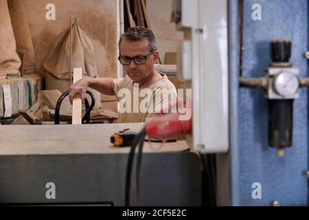 Operaio attento di mezza età in occhiali e vestiti casual messa a fuoco e utilizzando una macchina elettrica mentre si lavora con il legno in luce moderno monolocale con carpenteria Foto Stock