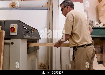 Vista laterale ad angolo basso di un artigiano medio attento al raccolto in occhiali e vestiti casual mettendo a fuoco e utilizzando carpenteria elettrica macchina mentre piallando legname in studio moderno chiaro Foto Stock