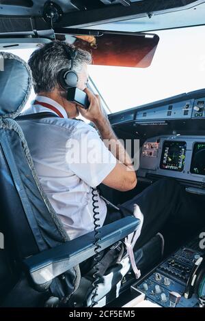 Vista laterale del capitano maschio che comunica tramite radio VHF mentre seduto in cabina di pilotaggio di aerei moderni e in preparazione per il volo Foto Stock