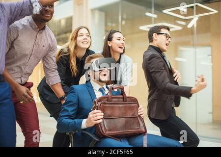 Il team del gruppo Business People è riuscito a spingere un collega indossando un visore per la realtà virtuale seduto in poltrona, i giovani uomini d'affari sorridono alzati le mani Foto Stock