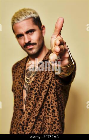 Ragazzo adulta con un taglio elegante e un tatuaggio vestito camicia leopardata che fa il movimento della pistola del dito e che guarda la macchina fotografica su sfondo giallo Foto Stock