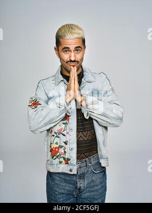 Giovane ragazzo alla moda in elegante giacca in denim che si accega con ripiegato mani vicino al mento e guardando la fotocamera su sfondo grigio Foto Stock