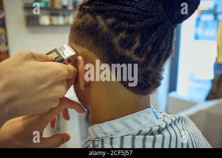 Ritagliare la vista da dietro di hairstylist anonimo facendo un moderno taglio di capelli con un rasoio ad un cliente afroamericano senza volto Foto Stock