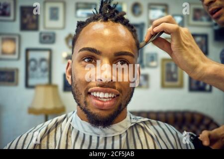 Ritagliare la vista di hairstylist anonimo facendo un taglio moderno a. un allegro cliente afro-americano Foto Stock