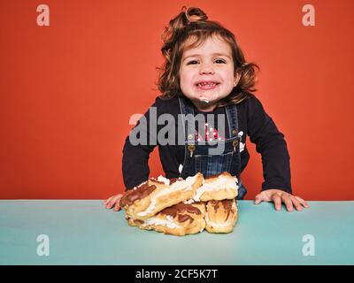 Carina bambina in abiti casual con il viso cosparso di crema bianca sorridente mentre si è in piedi al tavolo con un mucchio di eclair yummy contro parete rossa Foto Stock