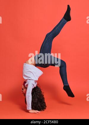 Vista laterale della ragazza teen in jeans e blusa che esegue supporto a mano su sfondo rosso in studio Foto Stock