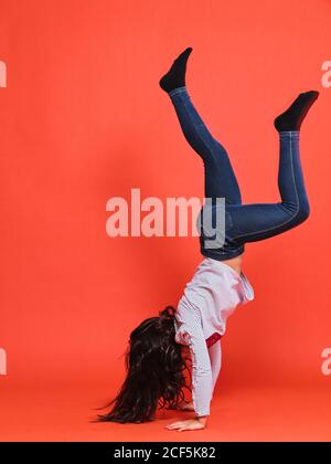 Vista laterale della ragazza teen in jeans e blusa che esegue supporto a mano su sfondo rosso in studio Foto Stock