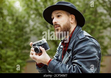 Giovane uomo bearded bello in cappello nero e giacca in pelle scattare foto in una strada verde Foto Stock