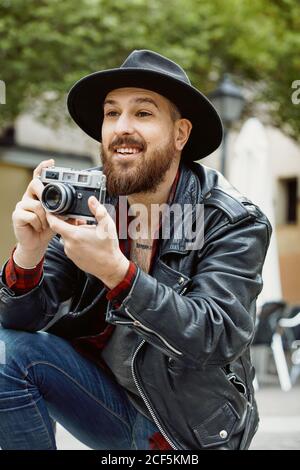 Giovane uomo bearded bello in cappello nero e giacca in pelle scattare foto in una strada verde Foto Stock