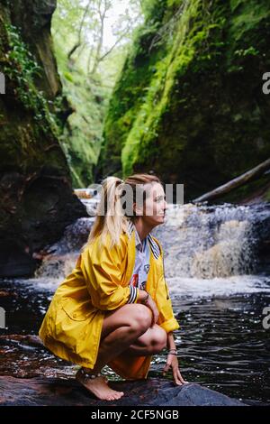 Vista laterale di una donna felice in giacca gialla accovacciata giù e toccante pietra vicino cascata passando tra le montagne in luce solare che guarda via Foto Stock