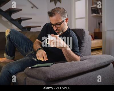 Uomo rilassato navigando su internet con un caffè da solo il divano nel soggiorno di casa Foto Stock