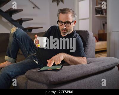 Uomo rilassato navigando su internet con un tè da solo il divano nel soggiorno di casa Foto Stock