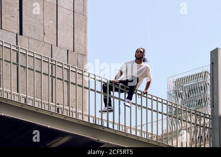 Giovane afroamericano seduto sulla ringhiera di ponte mentre trascorrere del tempo sulla strada della città in una giornata di sole Foto Stock