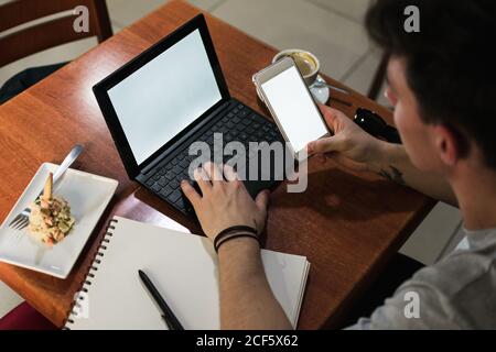 Da sopra Crop uomo seduto al tavolo e di navigazione portatile e smartphone con schermi vuoti mentre si lavora su progetti remoti in caffetteria Foto Stock